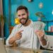 Smiling man at a desk points to a fan of dollar bills, sitting in front of a laptop in a modern room.