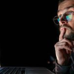 Man wearing glasses looks thoughtfully at a laptop screen displaying colorful financial charts and data.