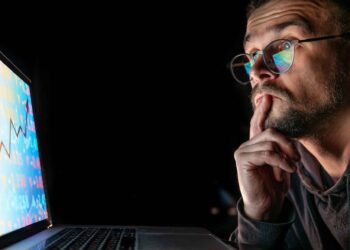 Man wearing glasses looks thoughtfully at a laptop screen displaying colorful financial charts and data.