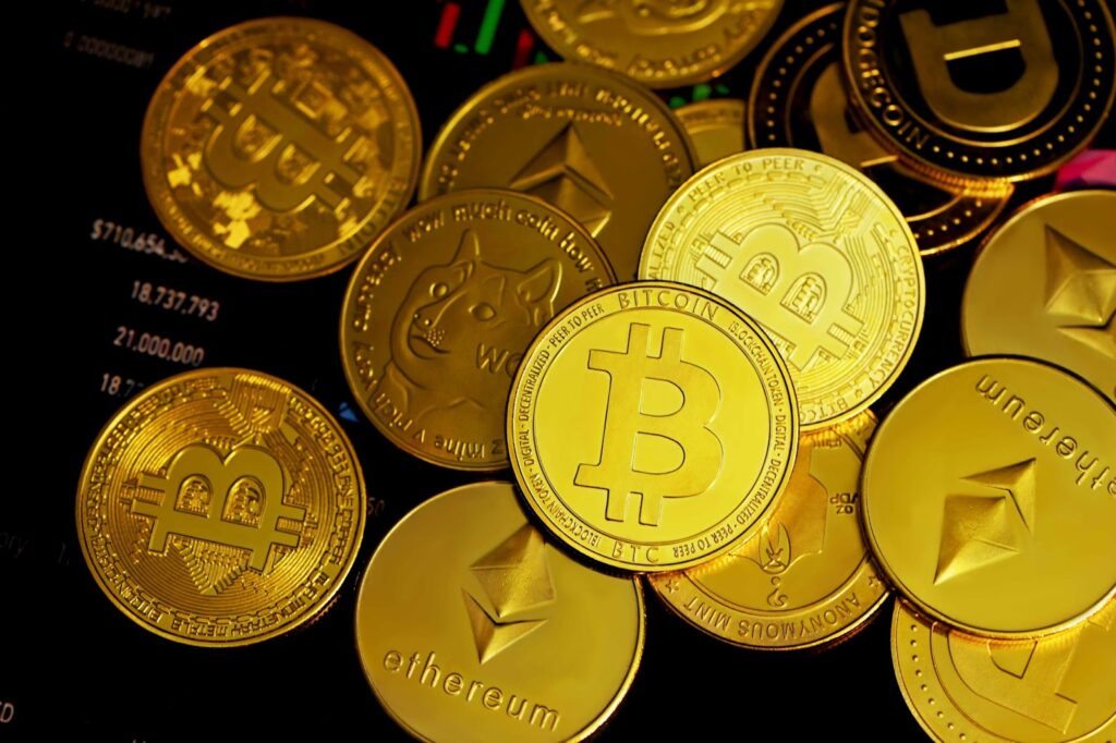 A pile of physical coins representing cryptocurrencies like Bitcoin, Ethereum, and Dogecoin sits atop a digital screen displaying financial data and charts, symbolizing the intersection of crypto assets and Venture Capital investments. | COYYN