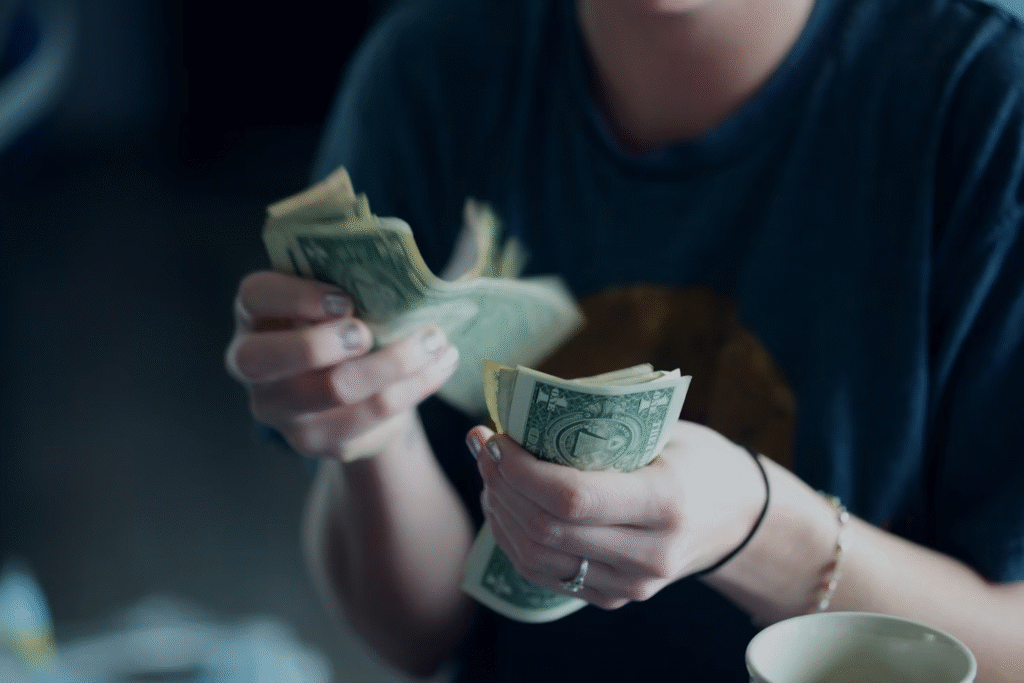 A person wearing a blue shirt is counting a stack of U.S. one-dollar bills, possibly from a personal loan, holding the cash in both hands. Their face is out of focus and partially out of frame. A white cup is visible in the lower right corner. | COYYN