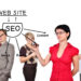 A woman in a red shirt writes "SEO" on a transparent board with arrows pointing to keywords like "Web Site," "Keyword," "Link," "Media," "Rank," "Page," and "Content." Three blurred people stand in the background. | COYYN