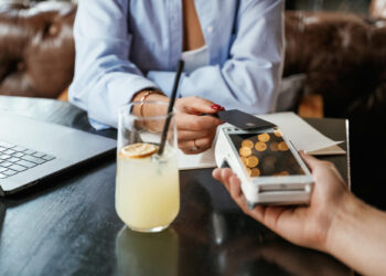 A person holds a credit card near a payment terminal for contactless payment at a table with a laptop, documents, and a glass of lemonade with ice and a lemon slice. | COYYN