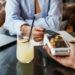 A person holds a credit card near a payment terminal for contactless payment at a table with a laptop, documents, and a glass of lemonade with ice and a lemon slice. | COYYN