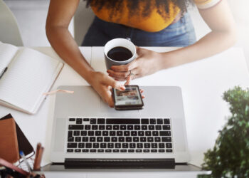 A person holding a cup of coffee and a smartphone sits at a desk with an open laptop, a notebook, and some stationery, viewed from above. | COYYN