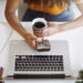 A person holding a cup of coffee and a smartphone sits at a desk with an open laptop, a notebook, and some stationery, viewed from above. | COYYN