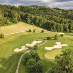 Aerial view of a golf course with neatly trimmed grass, winding pathways, multiple sand bunkers, scattered trees, and a dense green forest in the background under a partly cloudy sky. | COYYN
