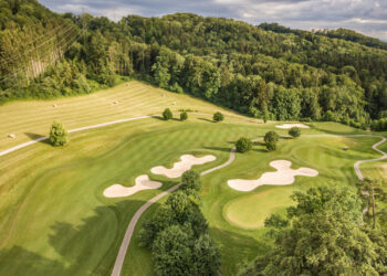 Aerial view of a golf course with neatly trimmed grass, winding pathways, multiple sand bunkers, scattered trees, and a dense green forest in the background under a partly cloudy sky. | COYYN