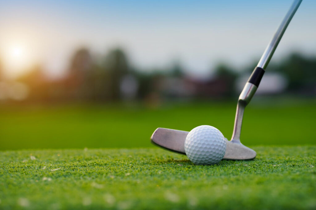 A close-up of a golf club about to hit a golf ball on green grass with a blurred background and the sun setting in the distance. | COYYN