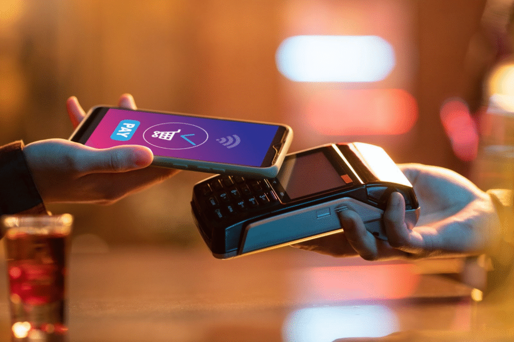 A person holds a smartphone displaying a P2P payment screen near a card reader held by another person, processing a contactless payment at a counter. | COYYN