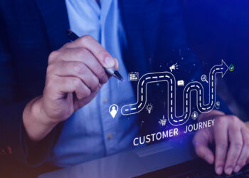 A person in a suit holds a pen, pointing at a digital illustration of a winding path labeled "Customer Journey" with icons for calendar, location, airplane, shopping cart, and communication along the route. | COYYN