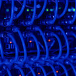 Close-up view of multiple blue network cables connected to server ports, with small indicator lights glowing green and orange in the background, creating a high-tech, data center atmosphere. | COYYN