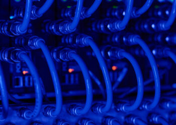 Close-up view of multiple blue network cables connected to server ports, with small indicator lights glowing green and orange in the background, creating a high-tech, data center atmosphere. | COYYN