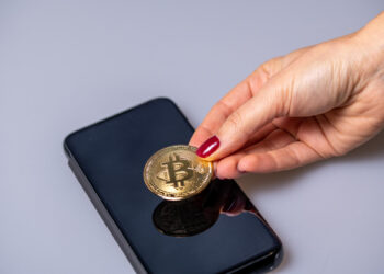 A hand with red nail polish places a gold Bitcoin coin on top of a black smartphone, both resting on a light gray surface. | COYYN