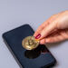 A hand with red nail polish places a gold Bitcoin coin on top of a black smartphone, both resting on a light gray surface. | COYYN