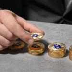 A person in a suit places a gold coin with a Bitcoin symbol onto one of several stacked piles of similar coins on a gray surface. | COYYN