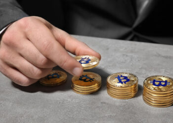 A person in a suit places a gold coin with a Bitcoin symbol onto one of several stacked piles of similar coins on a gray surface. | COYYN