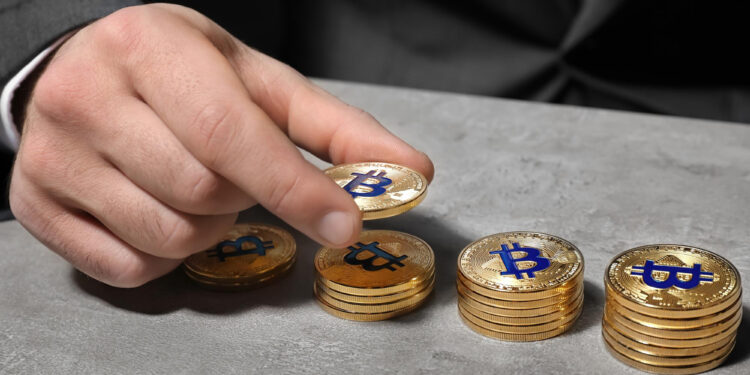 A person in a suit places a gold coin with a Bitcoin symbol onto one of several stacked piles of similar coins on a gray surface. | COYYN