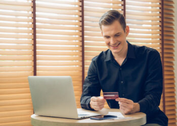 A smiling man in a black shirt sits at a table with a laptop, holding a credit card and looking at it. Papers and a smartphone are on the table, and wooden blinds cover the window behind him. | COYYN