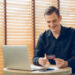 A smiling man in a black shirt sits at a table with a laptop, holding a credit card and looking at it. Papers and a smartphone are on the table, and wooden blinds cover the window behind him. | COYYN