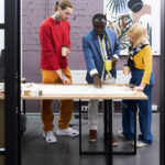 Three people stand around a desk in a modern office, discussing plans or blueprints. One person points at the paper, while the others hold coffee cups and listen attentively. Bright décor and plants are visible in the background. | COYYN