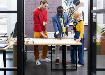 Three people stand around a desk in a modern office, discussing plans or blueprints. One person points at the paper, while the others hold coffee cups and listen attentively. Bright décor and plants are visible in the background. | COYYN