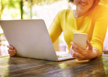 A woman in a yellow top sits at a wooden table using a laptop and holding a smartphone, smiling, with sunlight and greenery in the background. | COYYN