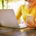 A woman in a yellow top sits at a wooden table using a laptop and holding a smartphone, smiling, with sunlight and greenery in the background. | COYYN