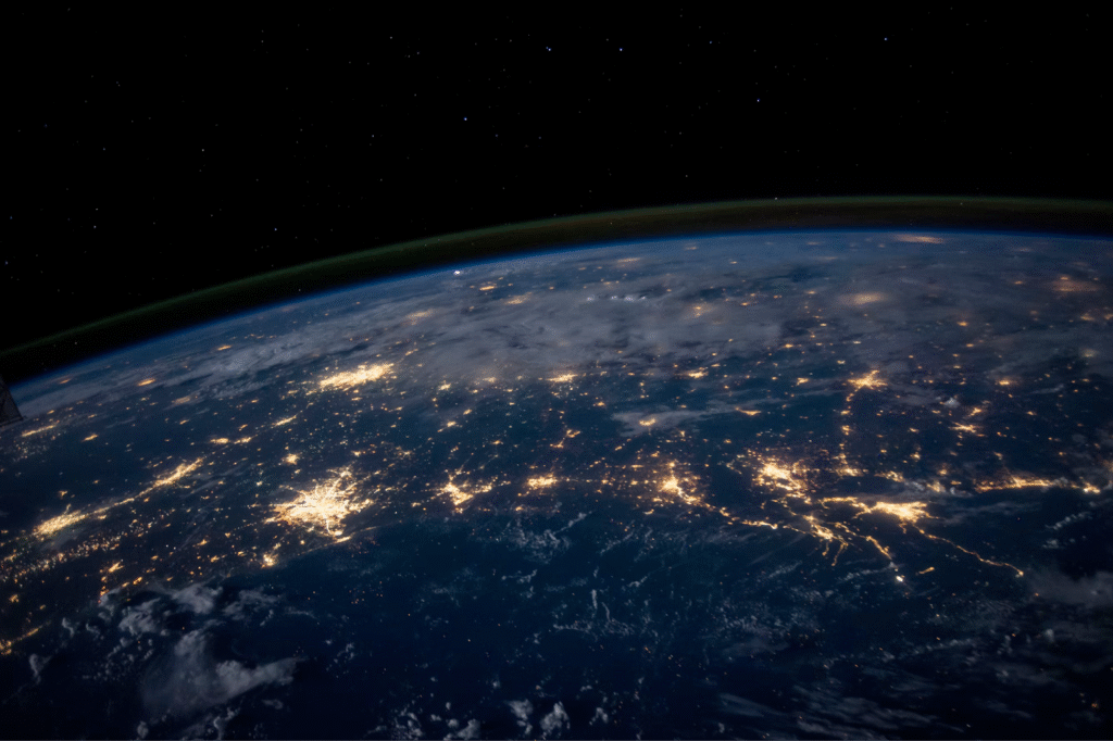 A view of Earth from space at night, showing illuminated city lights scattered across the surface, with a dark sky and a faint glow along the planet's curved horizon. | COYYN