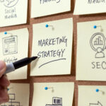 A hand writes “MARKETING STRATEGY” on a sticky note pinned to a corkboard, surrounded by other notes labeled “SEO,” “Website,” and “Social Media,” representing business planning concepts. | COYYN