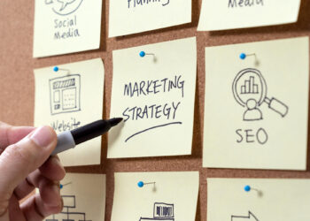 A hand writes “MARKETING STRATEGY” on a sticky note pinned to a corkboard, surrounded by other notes labeled “SEO,” “Website,” and “Social Media,” representing business planning concepts. | COYYN