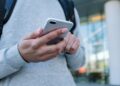 A person wearing a gray sweatshirt and backpack holds and uses a smartphone outdoors in front of a modern building with glass windows and a white pillar. | COYYN