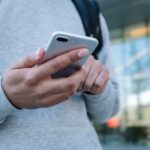 A person wearing a gray sweatshirt and backpack holds and uses a smartphone outdoors in front of a modern building with glass windows and a white pillar. | COYYN