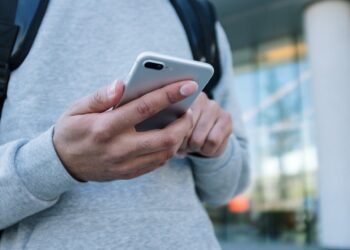 A person wearing a gray sweatshirt and backpack holds and uses a smartphone outdoors in front of a modern building with glass windows and a white pillar. | COYYN