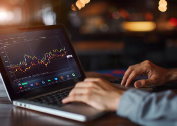A person uses a laptop displaying a colorful candlestick stock chart in a dimly lit setting, suggesting financial analysis or trading activity. | COYYN
