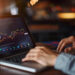 A person uses a laptop displaying a colorful candlestick stock chart in a dimly lit setting, suggesting financial analysis or trading activity. | COYYN