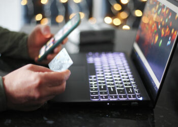 A person holds a smartphone in one hand and a credit card in the other while using a laptop with a lit keyboard. Out-of-focus lights create a festive background ambiance. | COYYN