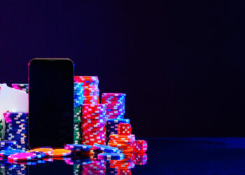 A smartphone stands among colorful poker chips, playing cards, and dice on a reflective surface, with a dark background, suggesting an online casino or gambling theme. | COYYN
