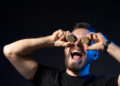 A man with a beard holds two Bitcoin coins over his eyes and smiles widely with his mouth open, wearing a black T-shirt and smartwatch, set against a dark background. | COYYN