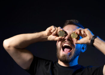A man with a beard holds two Bitcoin coins over his eyes and smiles widely with his mouth open, wearing a black T-shirt and smartwatch, set against a dark background. | COYYN
