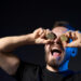 A man with a beard holds two Bitcoin coins over his eyes and smiles widely with his mouth open, wearing a black T-shirt and smartwatch, set against a dark background. | COYYN