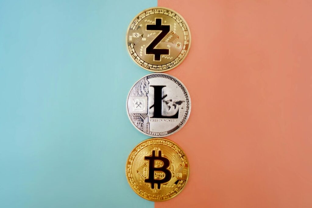 Three physical cryptocurrency coins are arranged vertically on a split background of peach and teal. From top to bottom, the coins display digital currency symbols for Zcash, Litecoin, and Bitcoin. | COYYN
