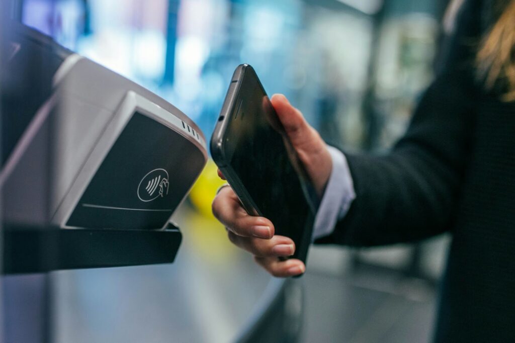 A person holds a smartphone close to a contactless payment terminal, making a digital payment. The background is blurred, focusing on the transaction. | COYYN