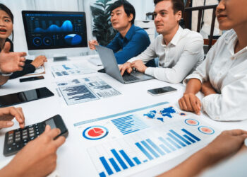 Five people sit around a table covered with charts, graphs, and electronic devices, having a business meeting. A monitor displays data visualizations in the background. | COYYN